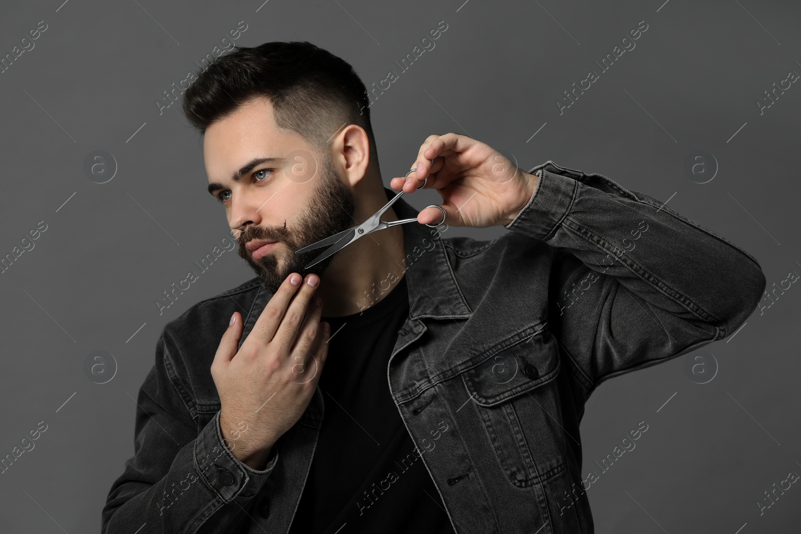 Photo of Handsome young man trimming beard with scissors on grey background