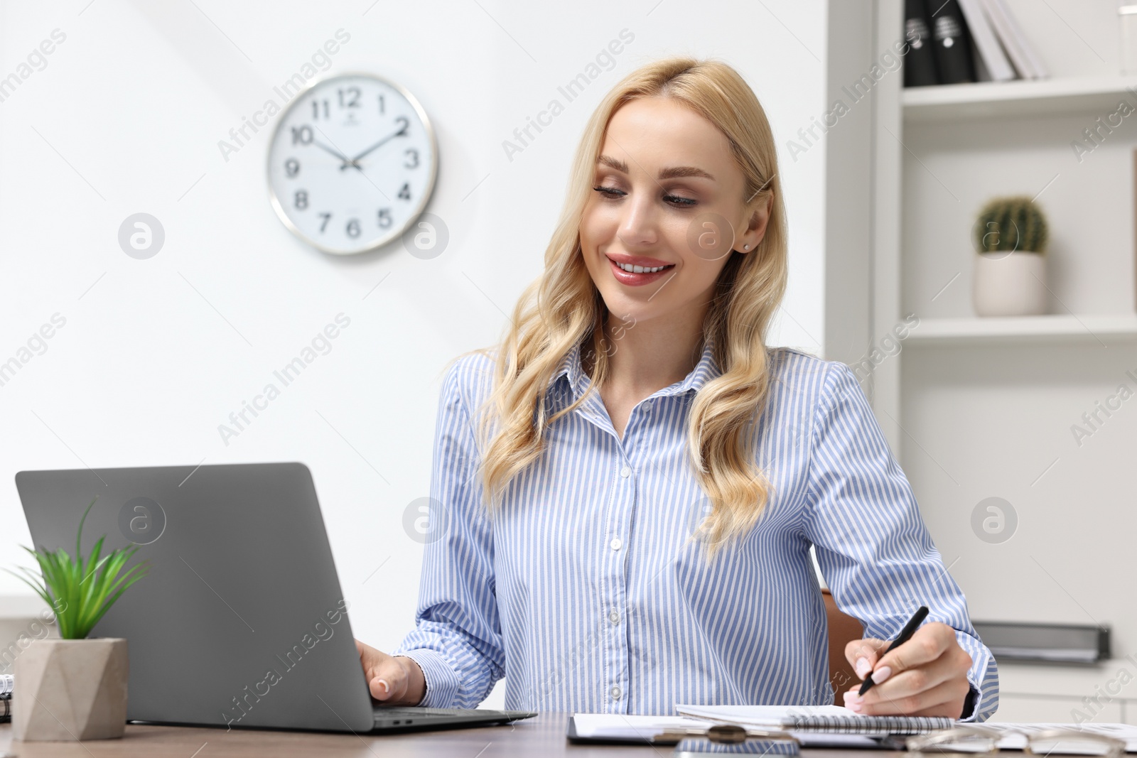Photo of Happy secretary taking notes while working with laptop at table in office