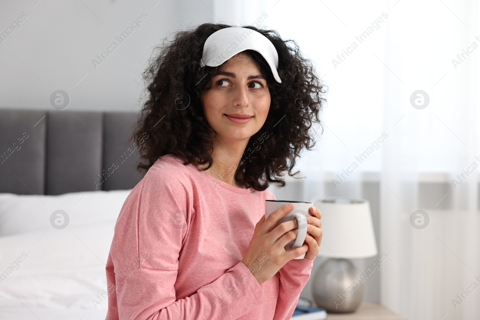 Photo of Beautiful young woman in stylish pyjama and sleep mask with cup of drink at home