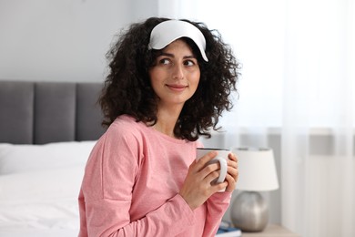 Photo of Beautiful young woman in stylish pyjama and sleep mask with cup of drink at home