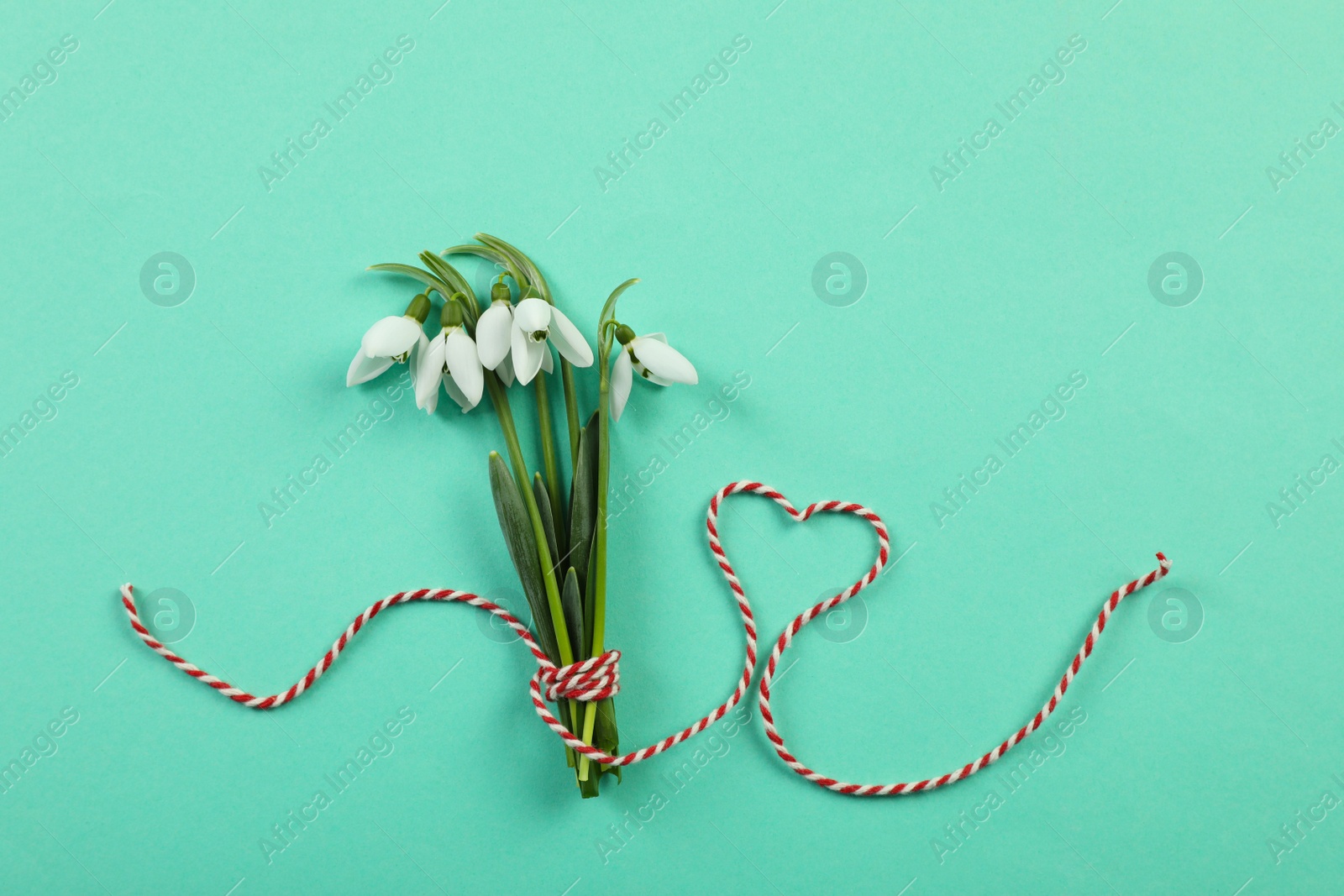 Photo of Beautiful snowdrops with traditional cord martisor on turquoise background, flat lay. Symbol of first spring day