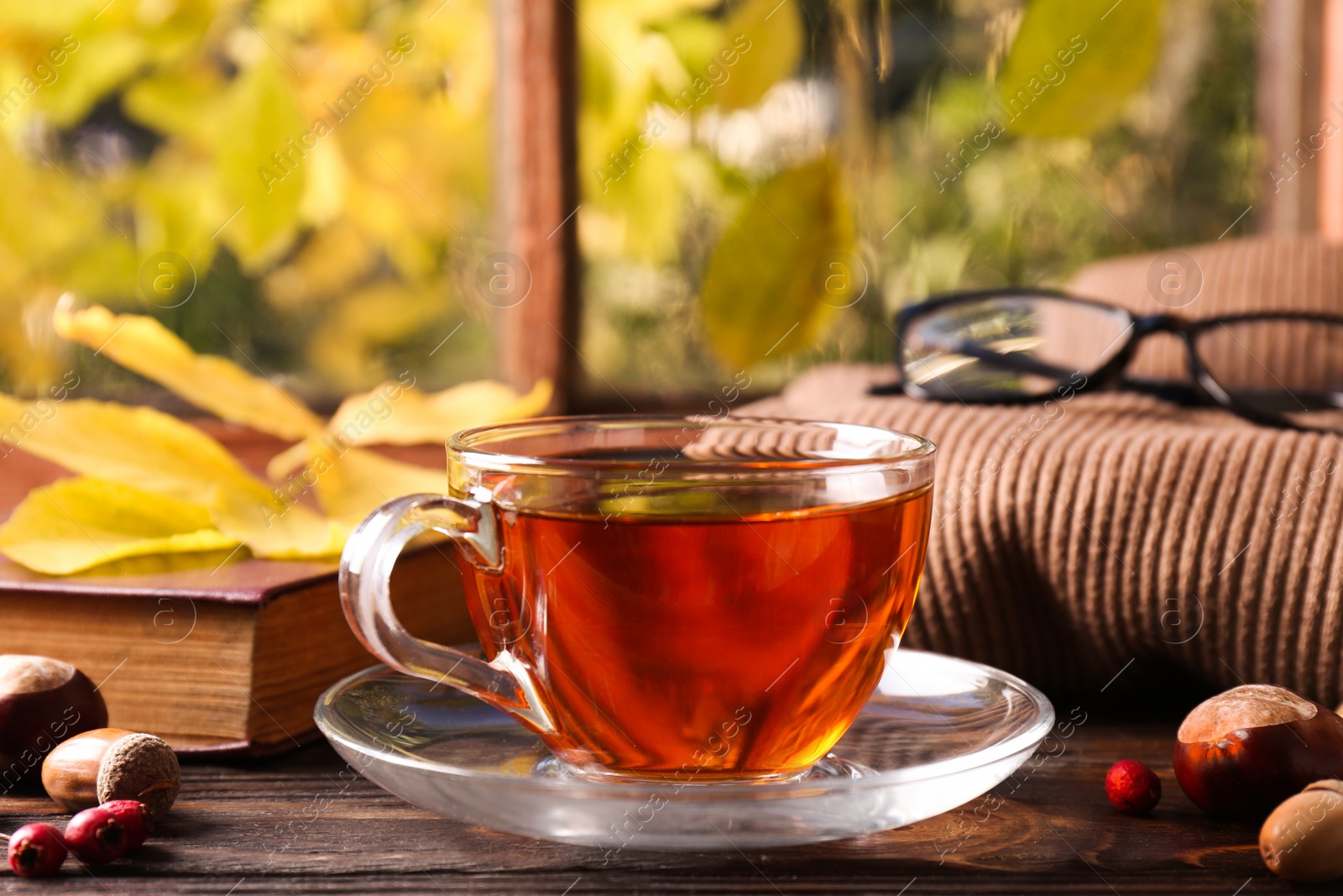 Photo of Cup of aromatic tea, acorns and soft sweater on wooden windowsill indoors. Autumn atmosphere