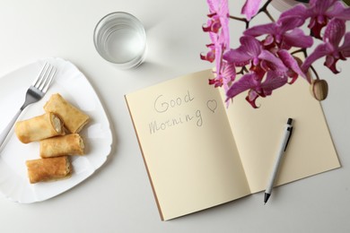 Notebook with inscription Good Morning, stuffed crepes and glass of water near beautiful blooming orchid on white table, flat lay