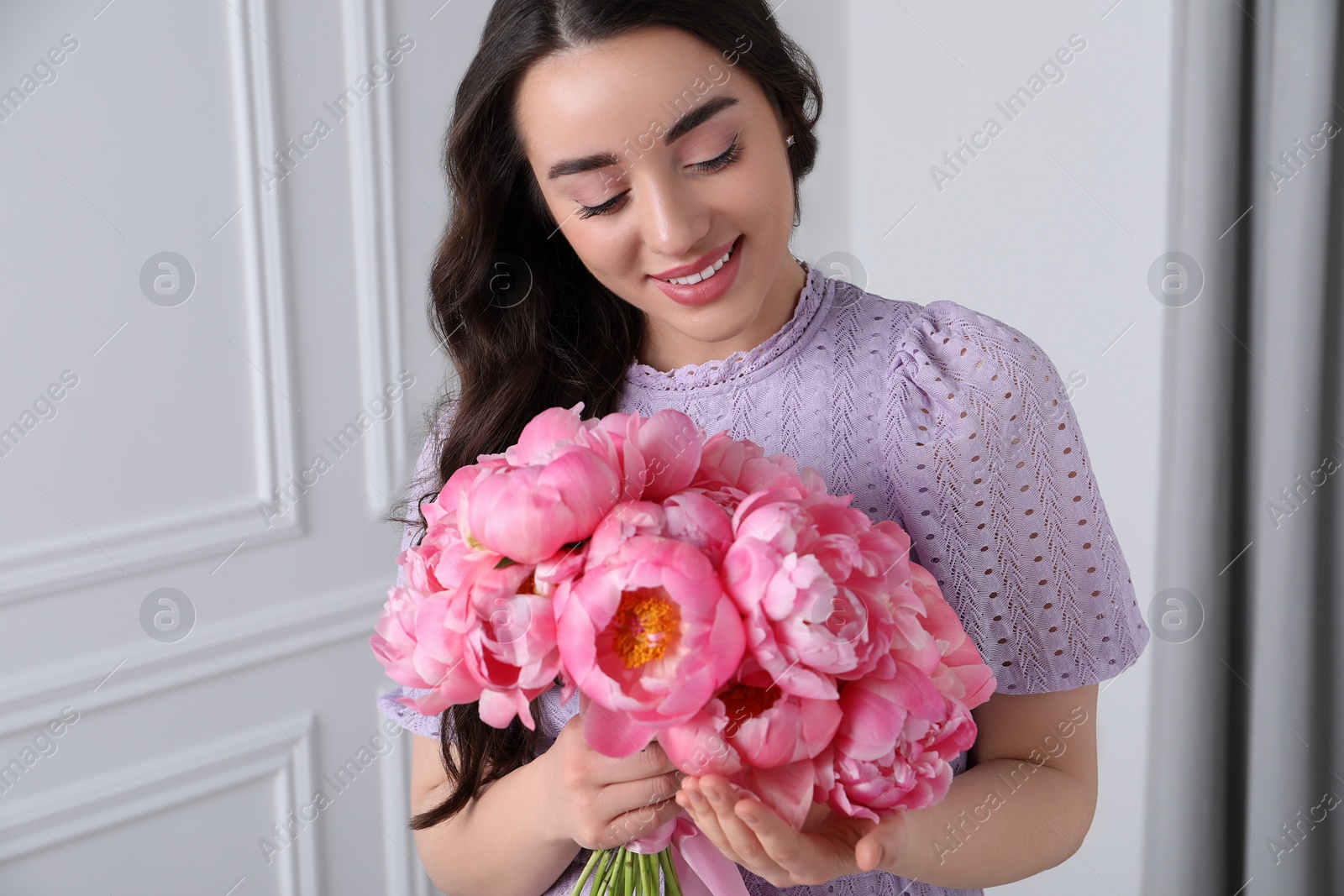 Photo of Beautiful young woman with bouquet of pink peonies indoors