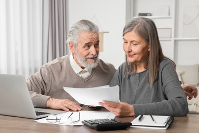 Elderly couple with papers and laptop discussing pension plan at wooden table in room