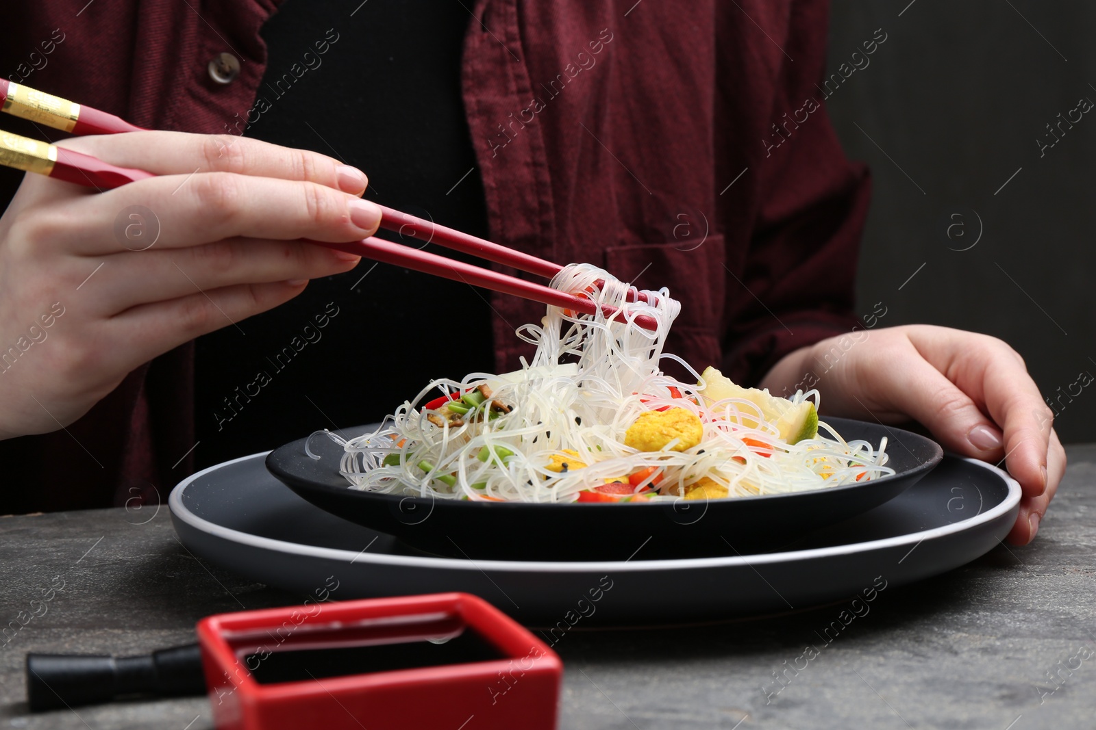 Photo of Stir-fry. Woman with chopsticks eating tasty rice noodles with meat and vegetables at grey textured table, closeup