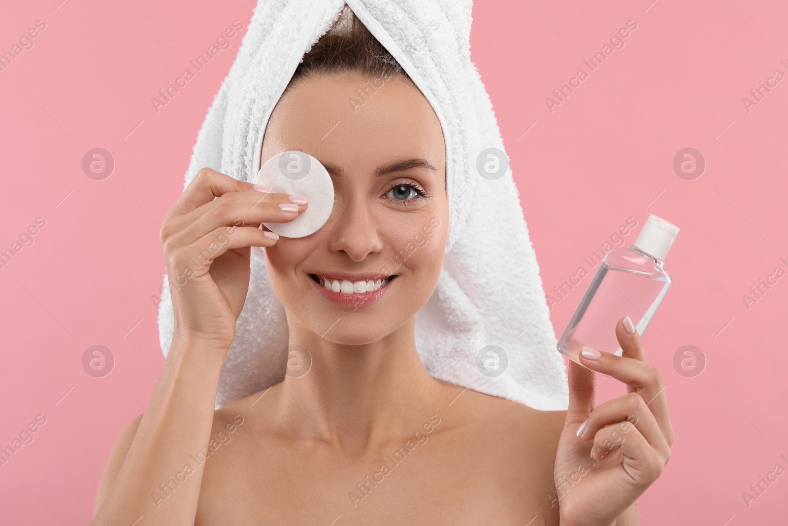 Photo of Smiling woman removing makeup with cotton pad and holding bottle on pink background