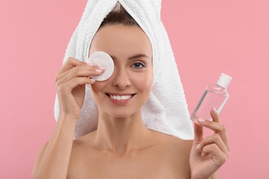 Smiling woman removing makeup with cotton pad and holding bottle on pink background