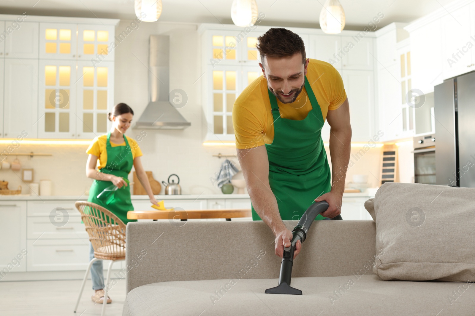 Photo of Professional janitor in uniform vacuuming sofa indoors