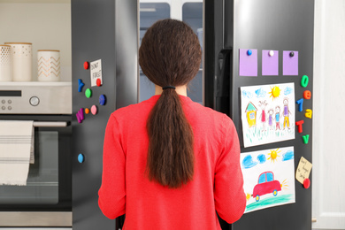 Photo of Woman opening refrigerator door with child's drawings, notes and magnets in kitchen