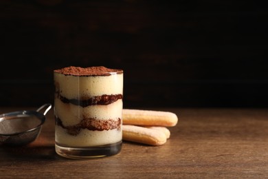 Photo of Delicious tiramisu in glass, biscuits and sieve on wooden table. Space for text