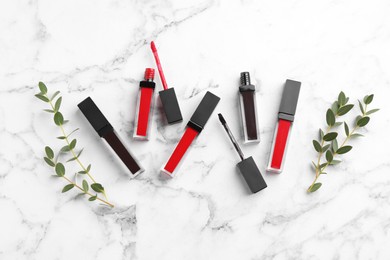 Photo of Different lip glosses, applicators and green leaves on white marble table, flat lay