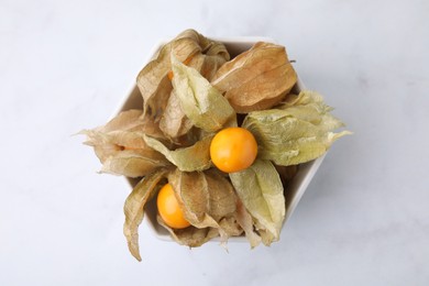 Ripe physalis fruits with calyxes in bowl on white marble table, top view