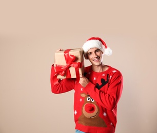 Photo of Happy man in Christmas sweater and Santa hat holding gift box on beige background