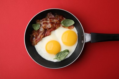 Fried eggs, bacon and basil in frying pan on red background, top view