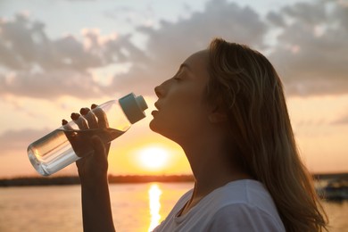 Photo of Young woman drinking water to prevent heat stroke near river at sunset