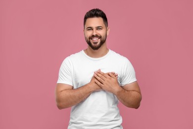 Photo of Thank you gesture. Handsome grateful man holding hands near heart pink background