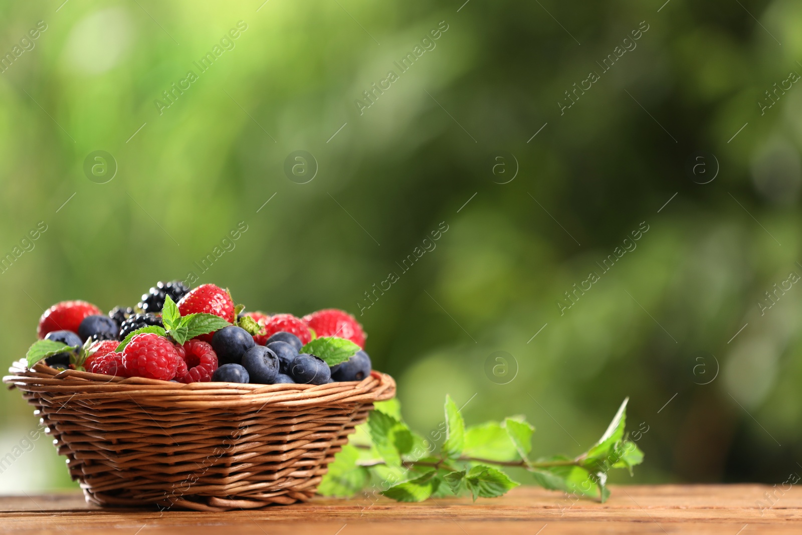 Photo of Wicker bowl with different fresh ripe berries and mint on wooden table outdoors, space for text