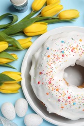 Photo of Delicious Easter cake decorated with sprinkles near beautiful tulips and painted eggs on light blue background, flat lay