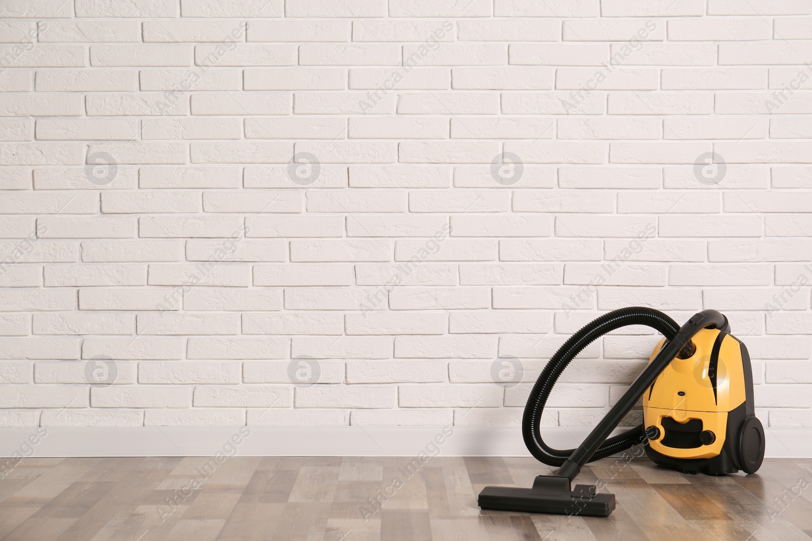 Photo of Modern yellow vacuum cleaner on wooden floor near white brick wall, space for text
