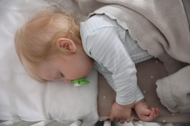 Photo of Cute little baby with pacifier sleeping in crib, top view. Bedtime