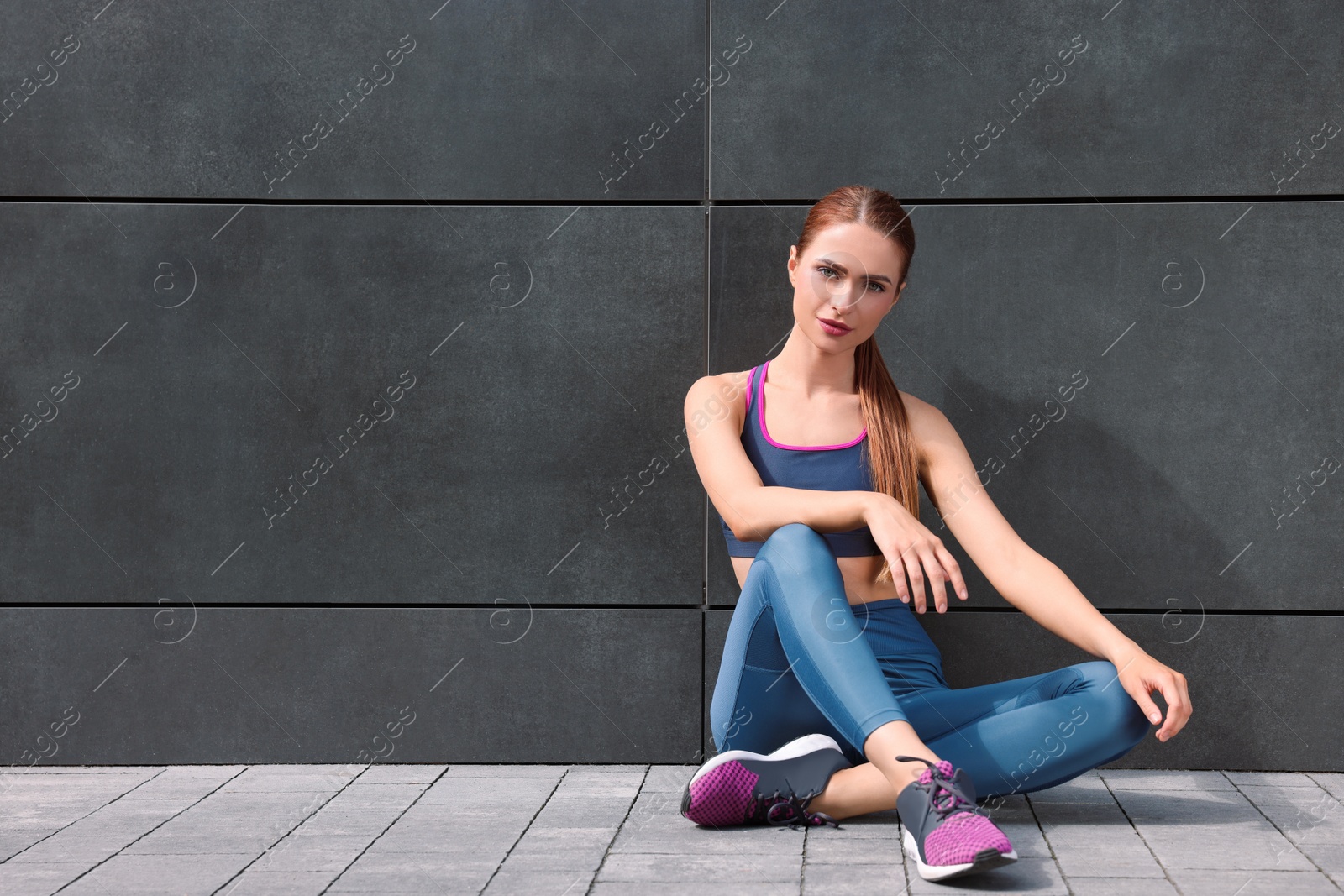 Photo of Beautiful woman in stylish gym clothes sitting near dark grey wall on street, space for text