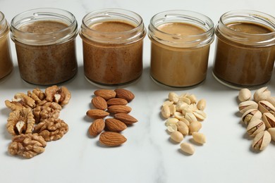 Photo of Tasty nut butters in jars and raw nuts on white marble table, closeup