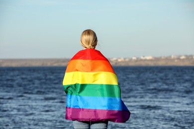 Photo of Woman wrapped in bright LGBT flag near river, back view