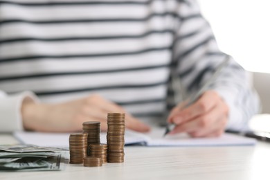 Financial savings. Woman making notes at white wooden table, focus on stacked coins