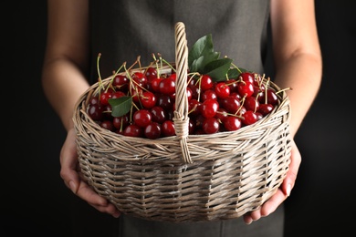 Photo of Woman holding wicker basket with sweet juicy cherries on black background, closeup