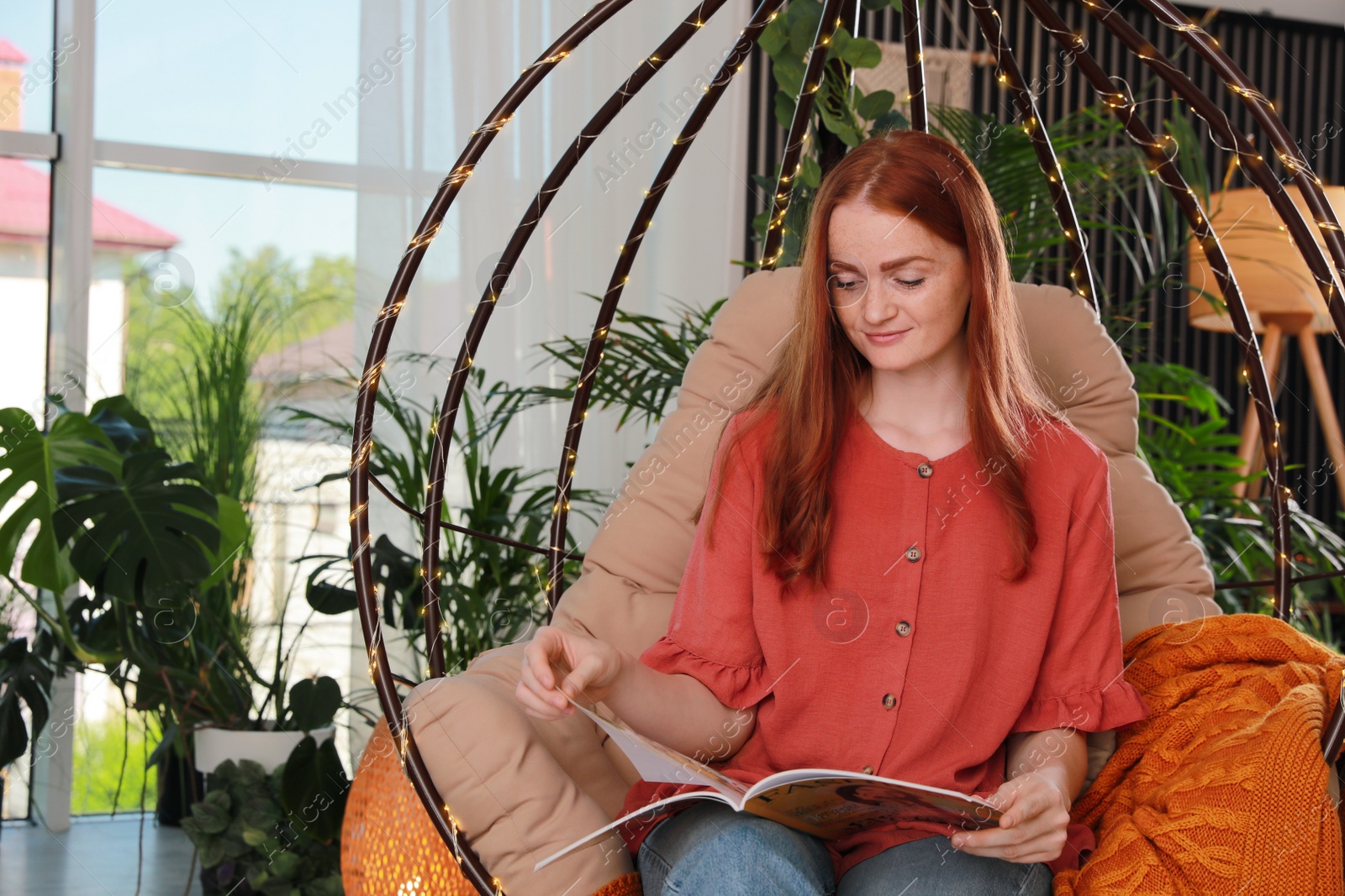 Photo of Happy young woman reading magazine in egg chair at indoor terrace