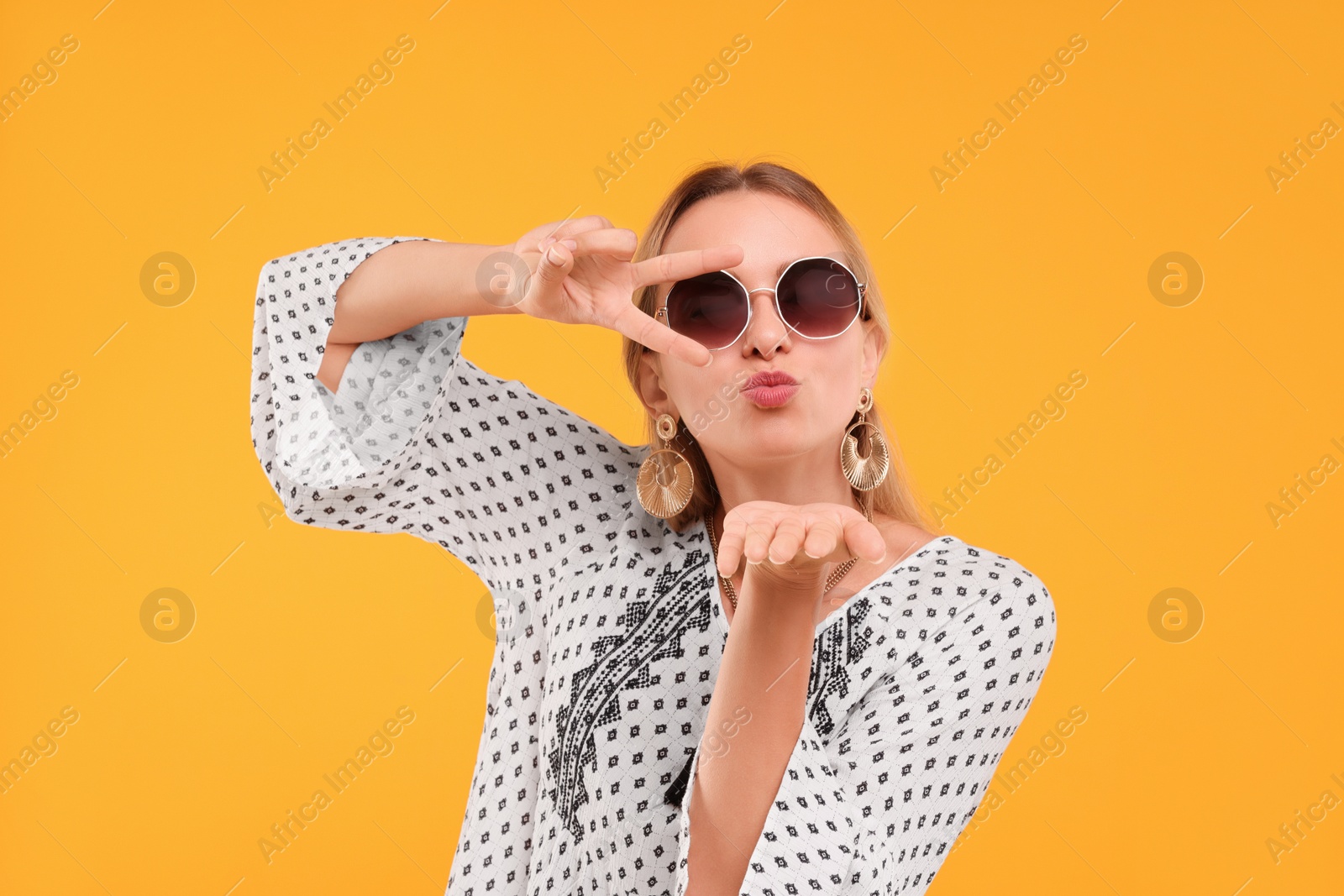 Photo of Portrait of beautiful hippie woman showing peace gesture and blowing kiss on yellow background