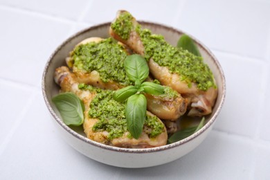Delicious fried chicken drumsticks with pesto sauce and basil in bowl on white tiled table, closeup