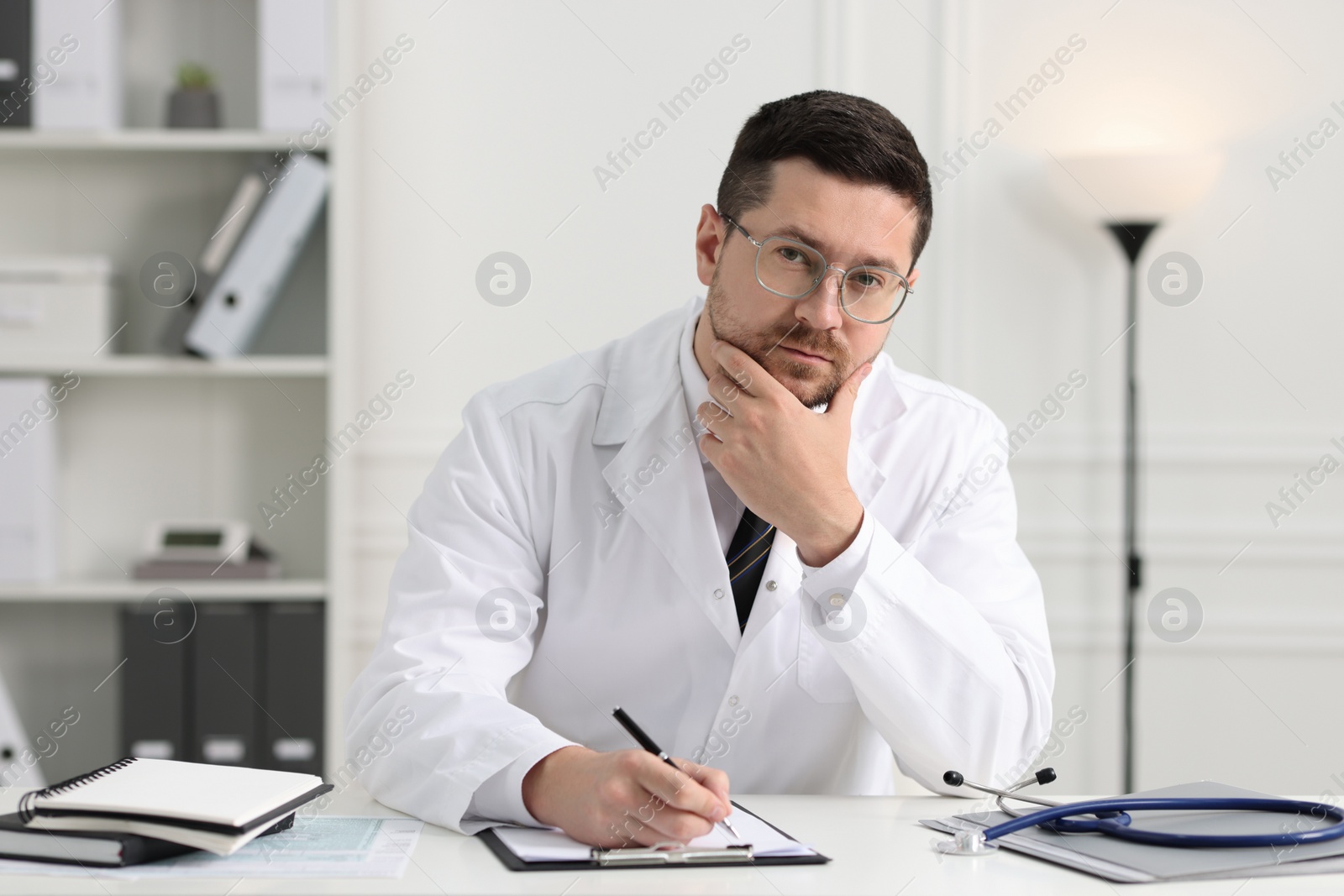 Photo of Doctor with pen and clipboard working at white table in clinic