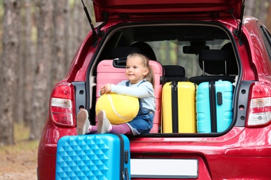 Photo of Cute little girl sitting in car trunk loaded with suitcases outdoors