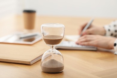 Photo of Hourglass with flowing sand on desk. Woman taking notes while using calculator indoors, selective focus