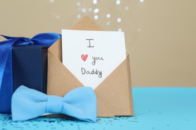 Photo of Happy Father's Day. Card with phrase I Love You, Daddy in envelope, bow tie and gift box on light blue table, closeup. Space for text