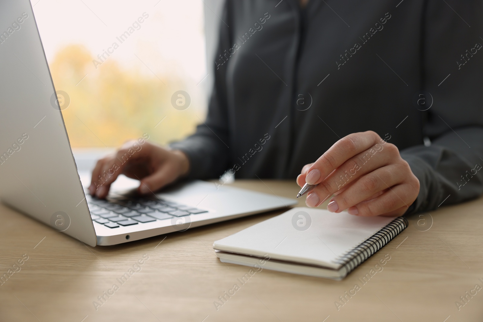 Photo of Woman with notebook and pen working on laptop at wooden table, closeup. Electronic document management