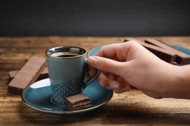 Woman with cup of delicious coffee and wafers at wooden table, closeup
