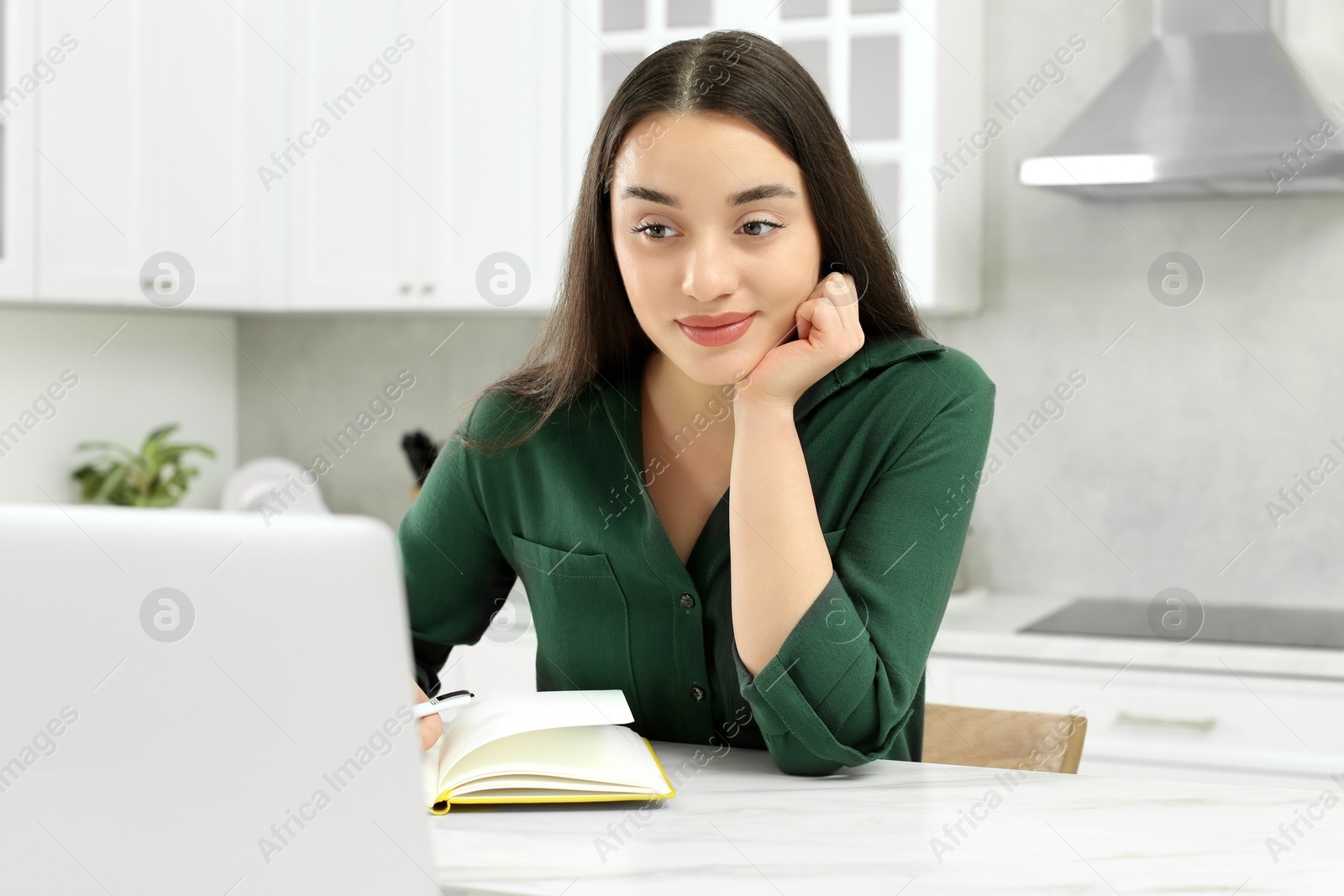 Photo of Home workplace. Woman with pen and notebook working on laptop at marble desk in kitchen