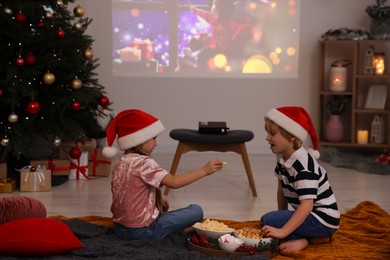 Photo of Brother and sister spending time together with snacks near video projector in room. Christmas atmosphere