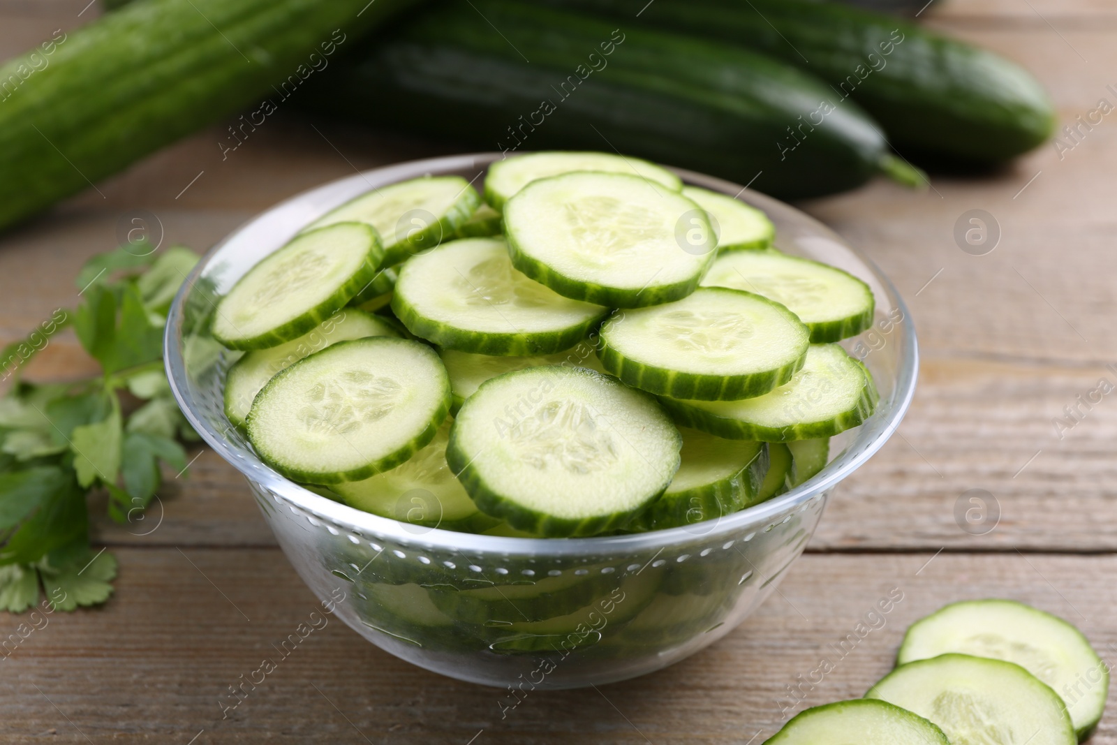 Photo of Cut cucumber in glass bowl, fresh vegetables and parsley on wooden table, closeup