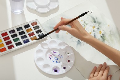 Woman painting flowers with watercolor at white table, above view. Creative artwork