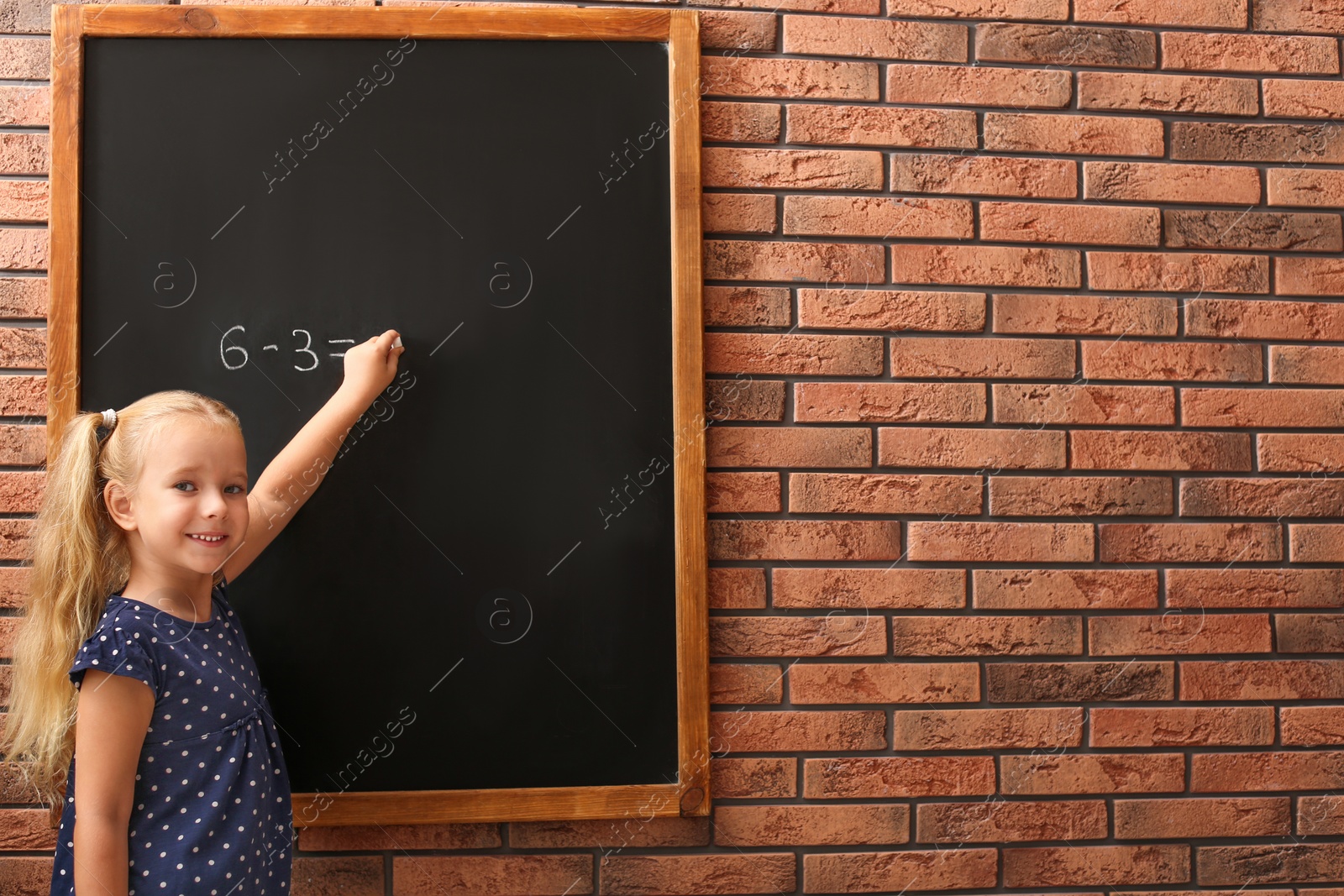 Photo of Cute little left-handed girl doing sums on chalkboard near brick wall