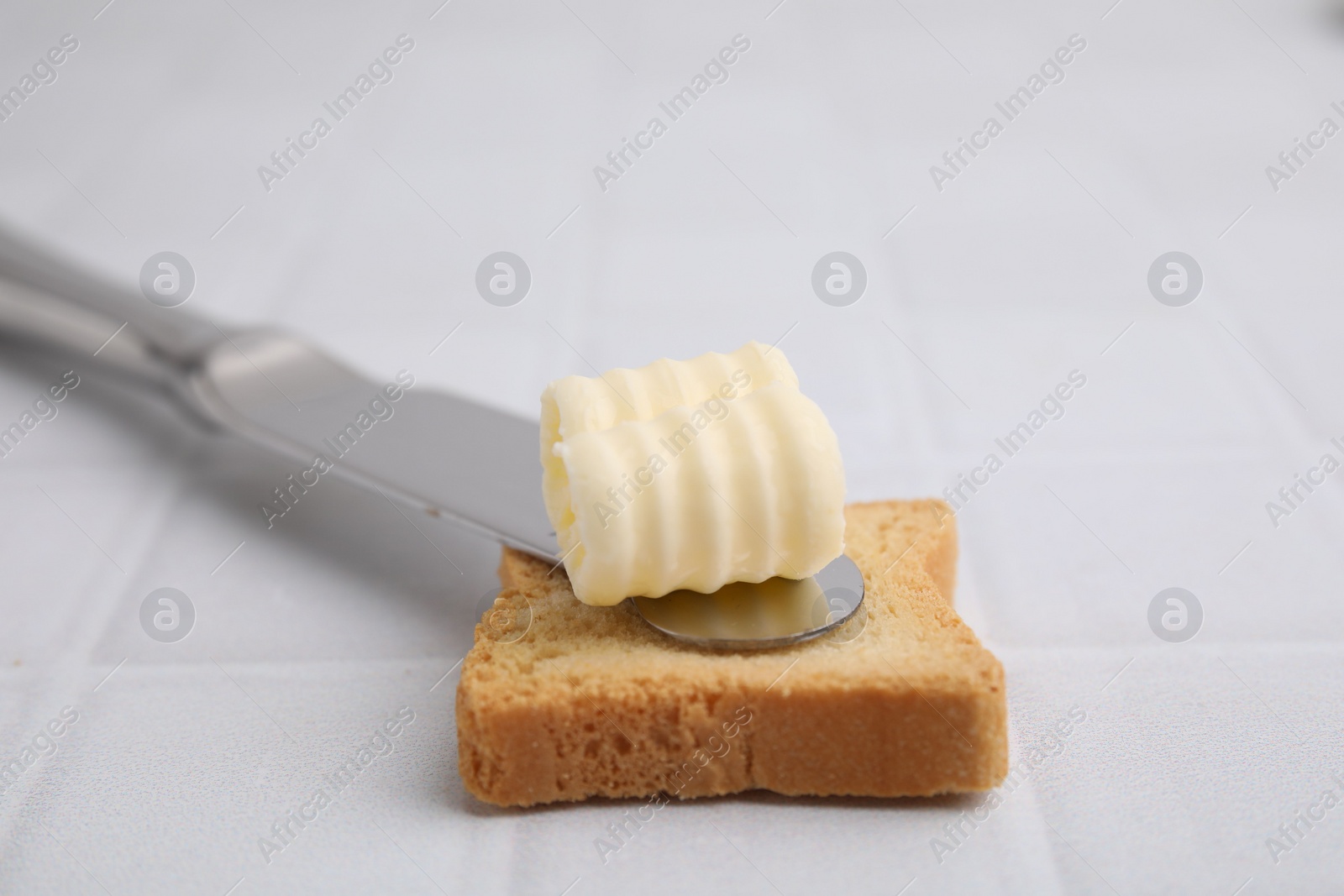 Photo of Tasty butter curl, knife and piece of dry bread on white tiled table, closeup