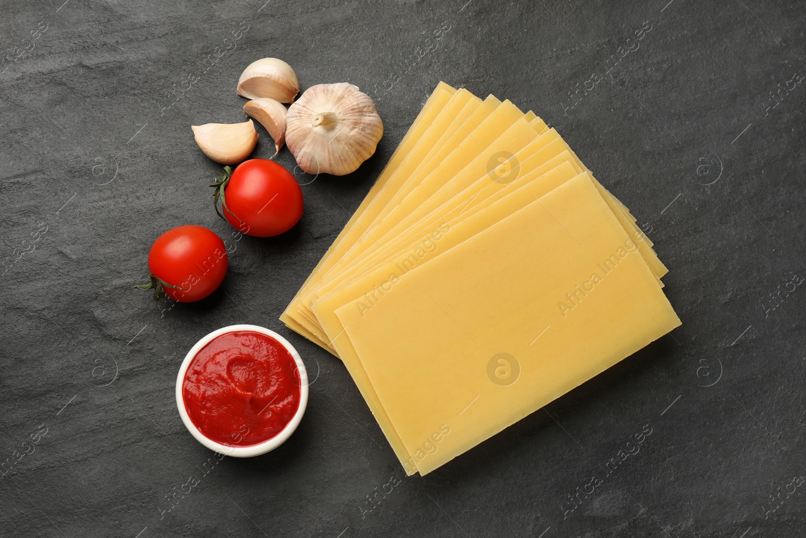 Photo of Cooking lasagna. Pasta sheets, tomato, garlic and ketchup on dark textured table, flat lay