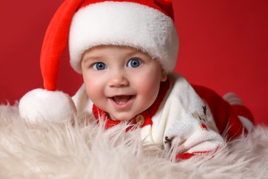 Cute baby in Santa hat on fluffy carpet against red background, closeup. Christmas celebration