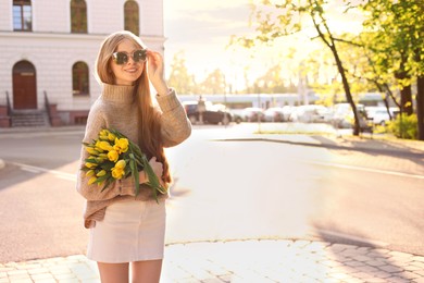 Beautiful teenage girl with bouquet of yellow tulips on city street, space for text