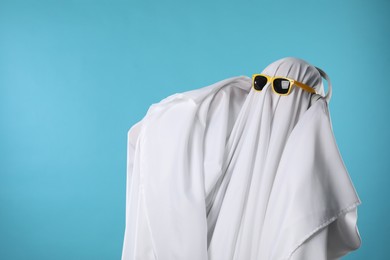 Stylish ghost. Person covered with white sheet in sunglasses and headphones on light blue background, space for text