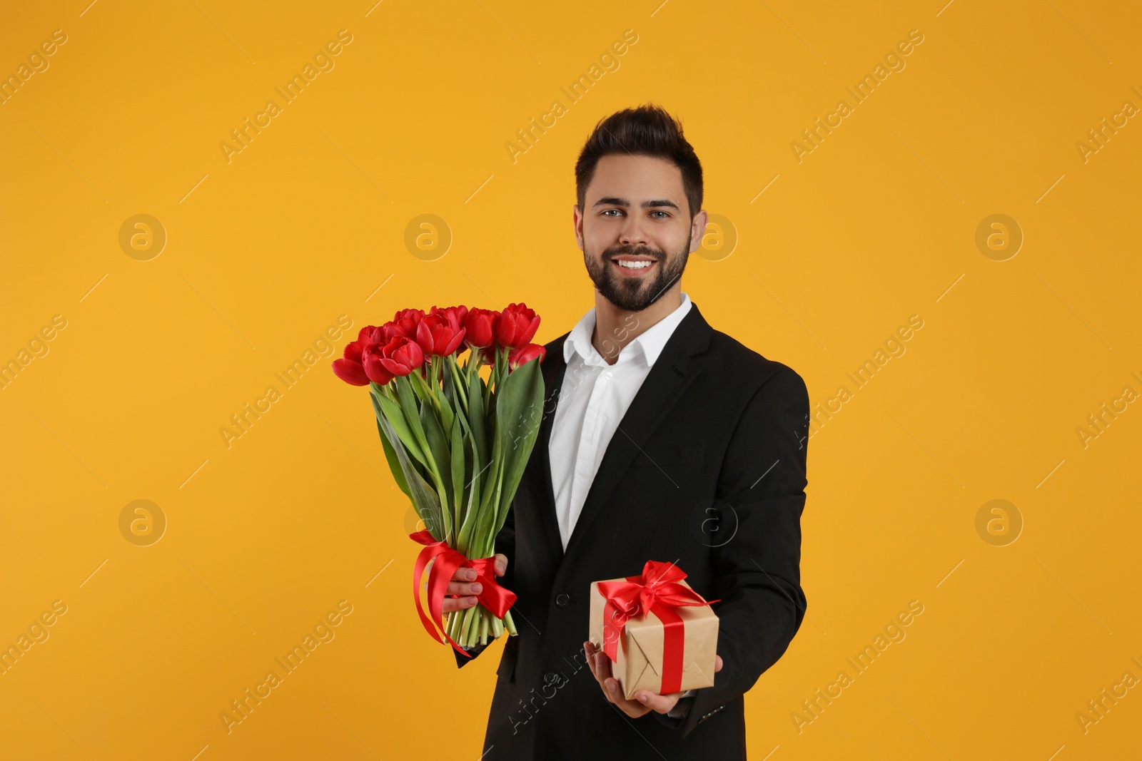 Photo of Happy man with red tulip bouquet and gift box on yellow background. 8th of March celebration
